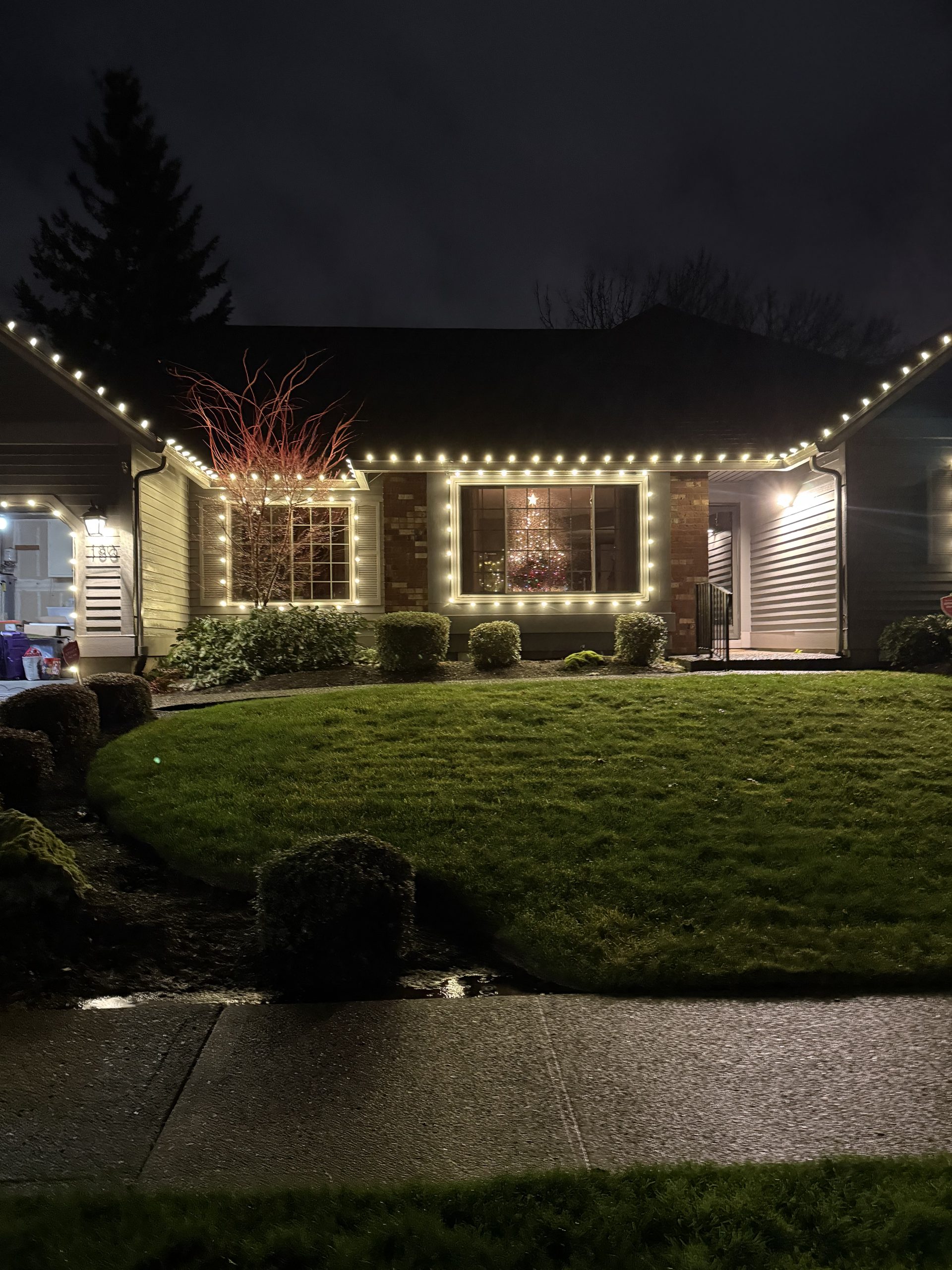 Single-story brick and siding home at night with warm white lights along the roof edges and framing two front windows